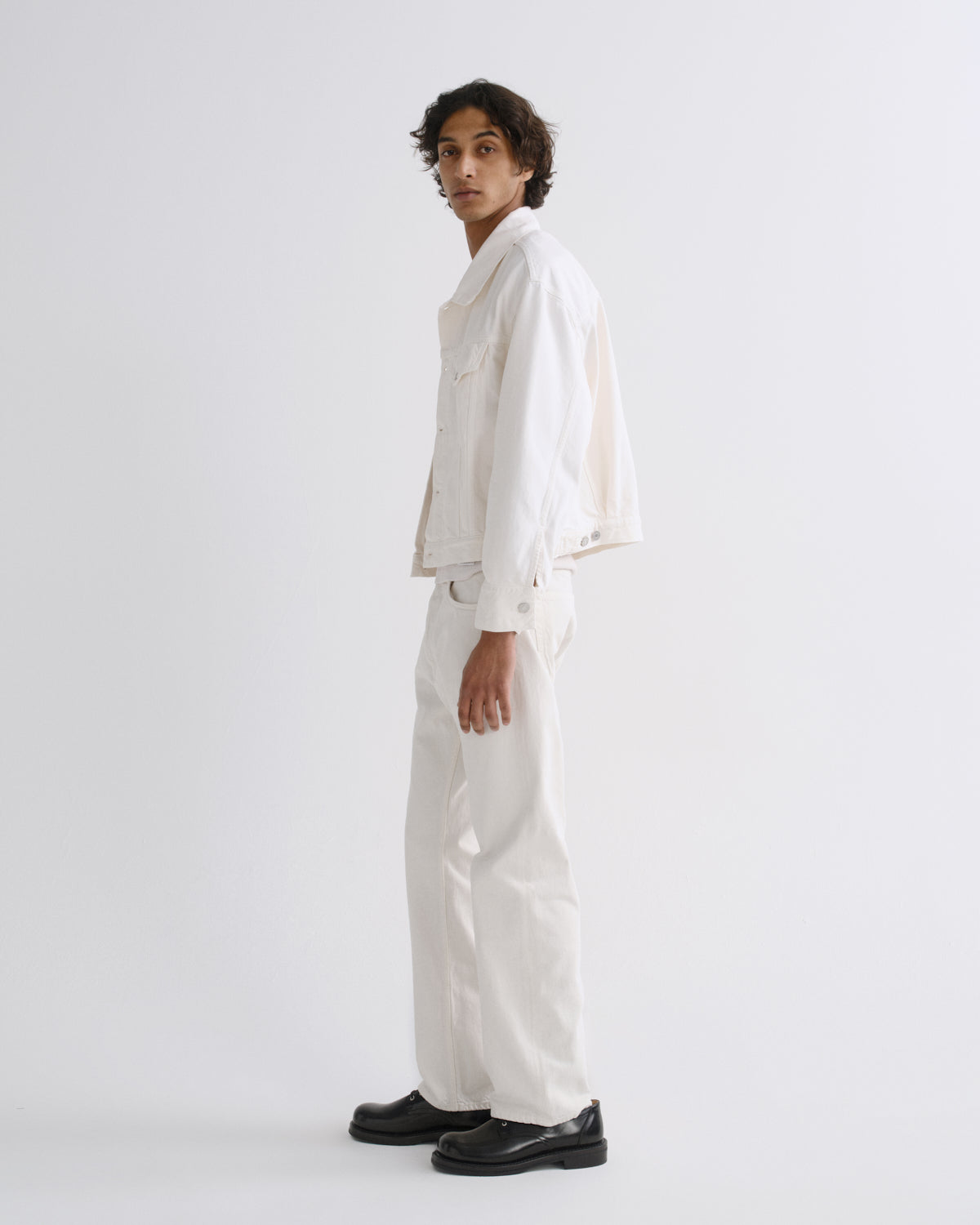 A model wearing a white cropped denim jacket, white wide-leg trousers, and black leather shoes, standing in profile against a light gray studio background.