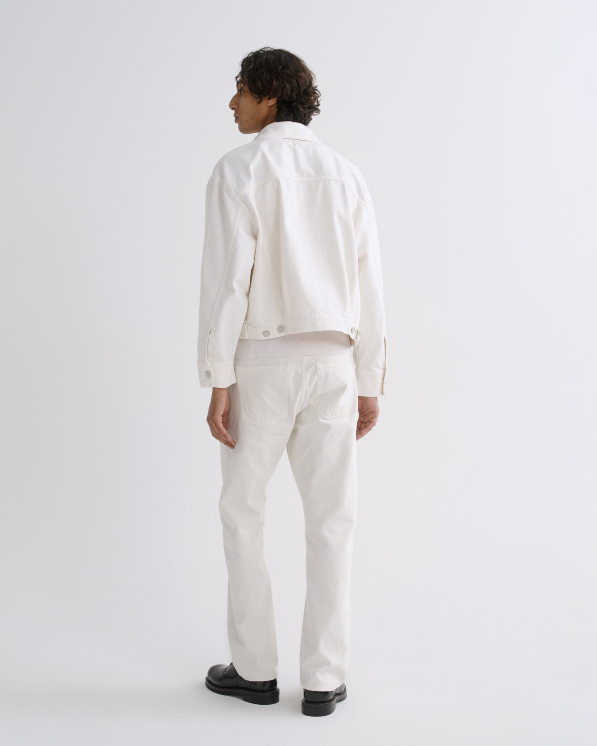 A model shown from the back wearing a white cropped denim jacket, white trousers, and black shoes against a plain white studio background.