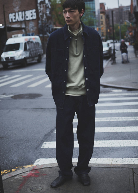 Man standing on a city street with a crosswalk, wearing a dark jacket and light shirt.