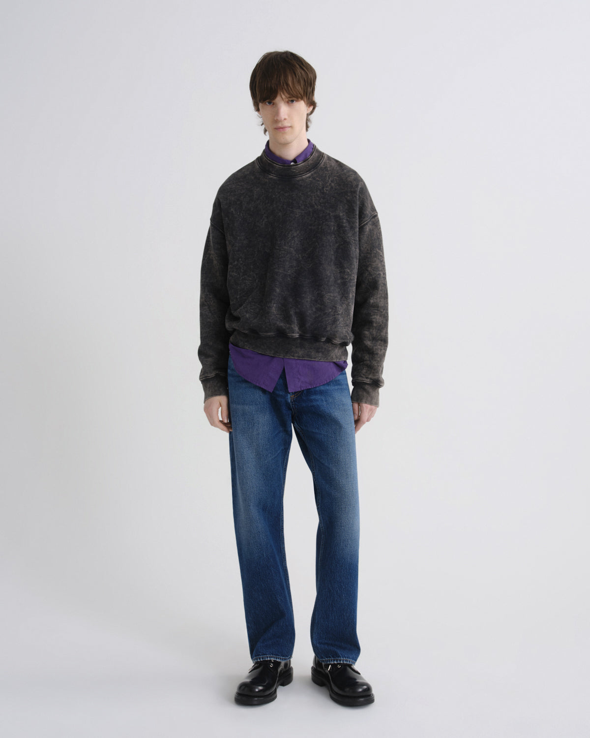 Person wearing a dark gray sweater, purple shirt, blue jeans, and black shoes on a white background