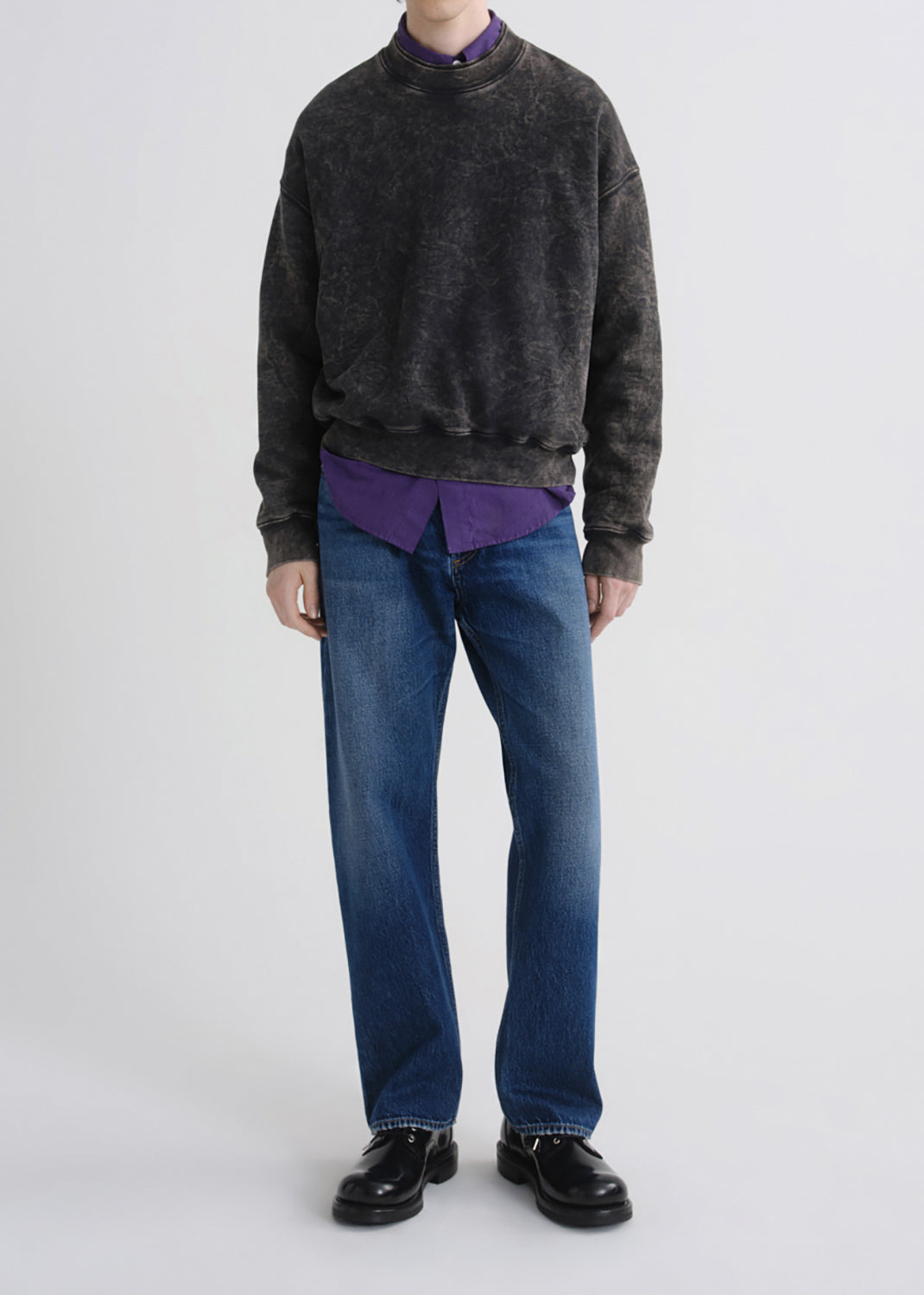 A model wearing a dark grey washed crewneck sweatshirt layered over a purple collared shirt, paired with blue jeans and black leather shoes, photographed against a grey background.