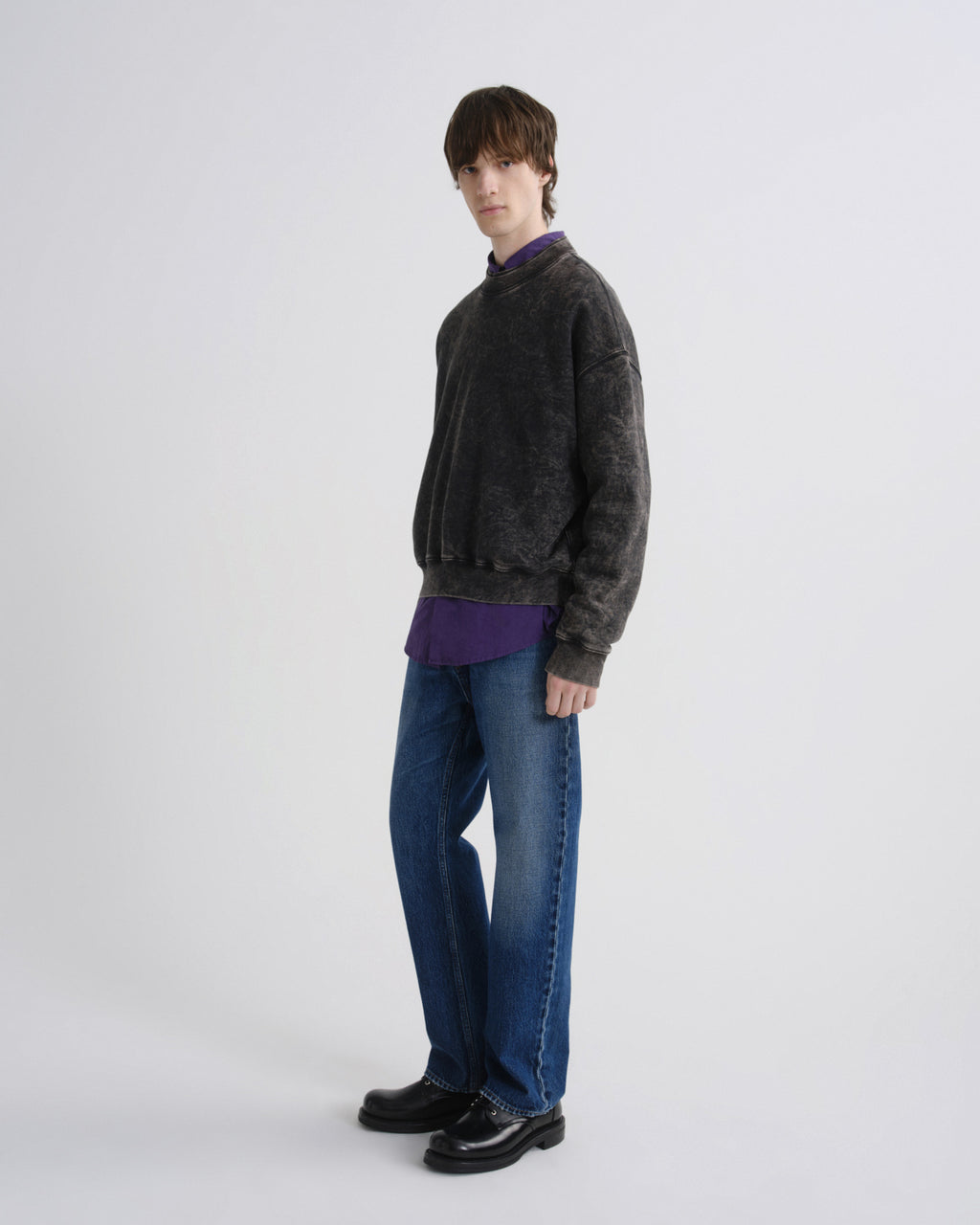 A model stands in profile wearing a dark charcoal gray washed crewneck sweater over a purple collared shirt, blue jeans, and black leather shoes, against a light gray studio background.