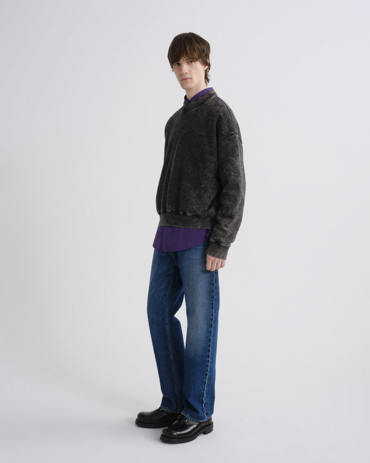 A model stands in profile wearing a dark charcoal gray washed crewneck sweater over a purple collared shirt, blue jeans, and black leather shoes, against a light gray studio background.