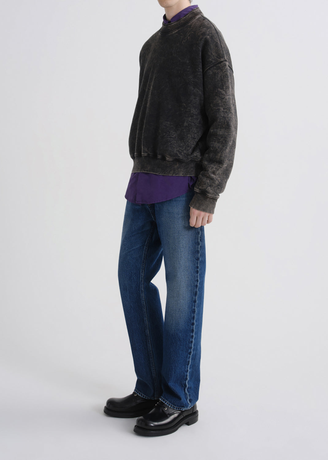 A model shown from the neck down wears a dark charcoal mineral-wash crewneck sweatshirt over a purple shirt, paired with blue jeans and black leather shoes, against a grey background.
