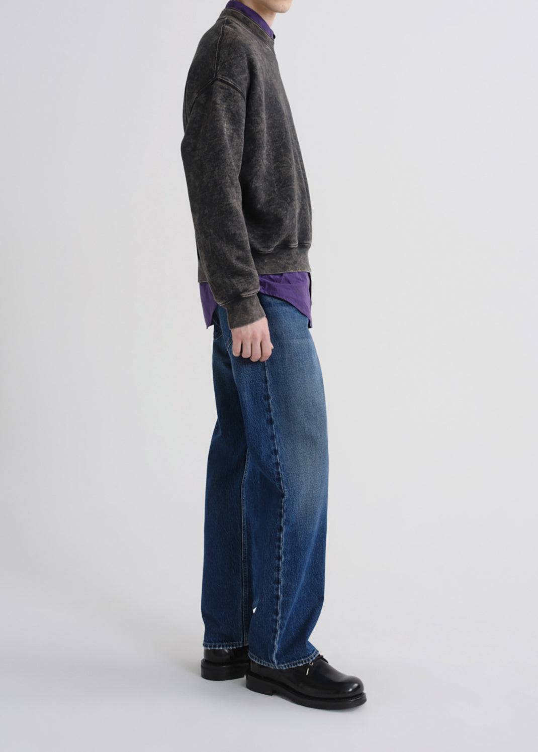 A side-profile view of a person wearing a dark grey knit sweater, a purple shirt underneath, wide-leg blue jeans, and black leather loafers, against a grey background.