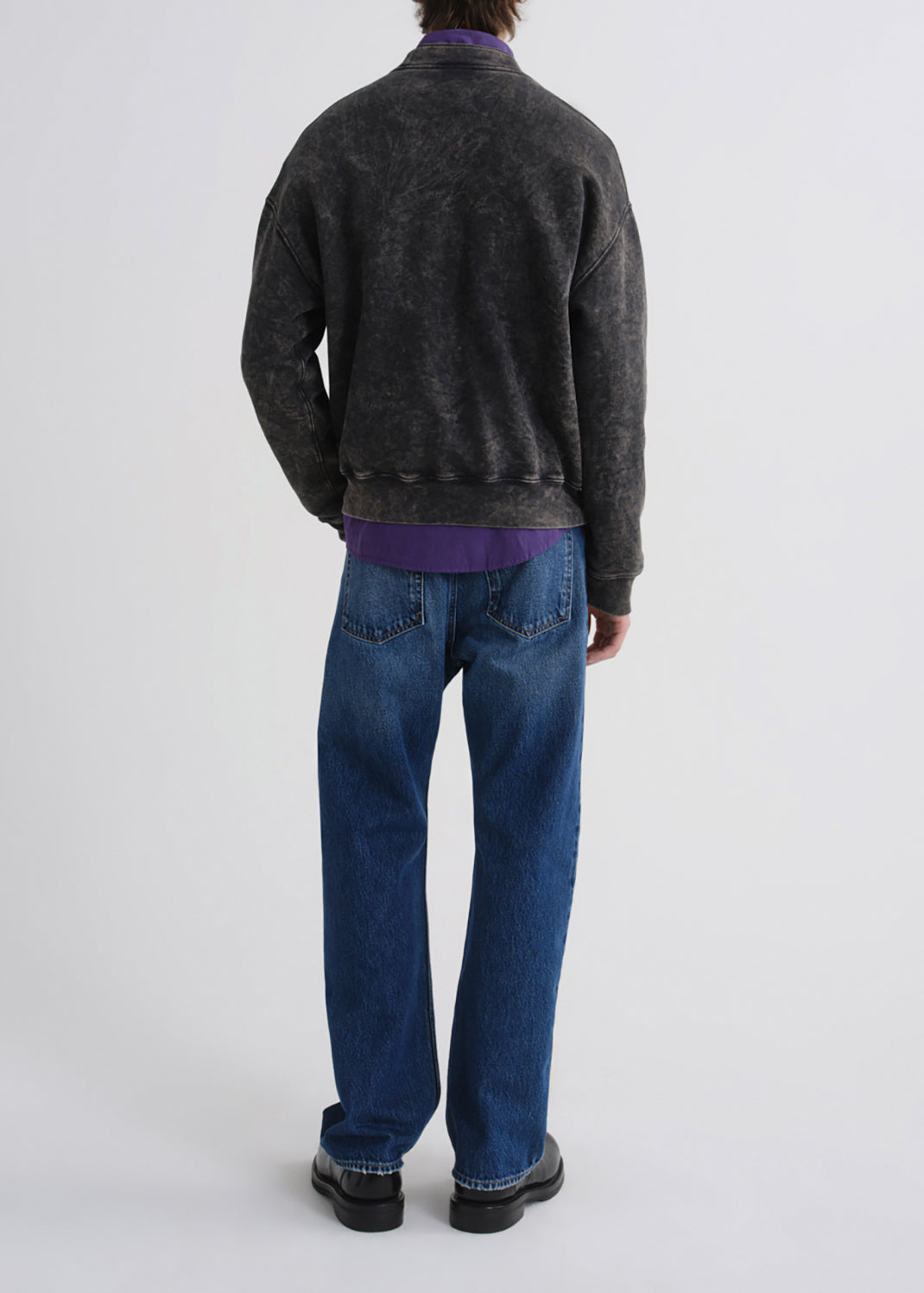 A person shown from the back wearing a dark charcoal washed bomber jacket over a purple shirt, blue jeans, and black shoes, photographed against a grey background.