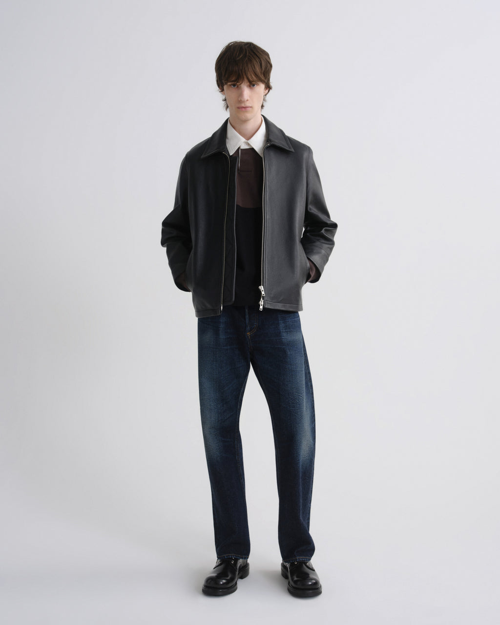 A male model stands against a light gray studio background wearing a dark gray leather jacket over a white collared shirt, dark brown sweater, blue jeans, and black shoes.