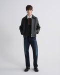 A male model stands against a light gray studio background wearing a dark gray leather jacket over a white collared shirt, dark brown sweater, blue jeans, and black shoes.