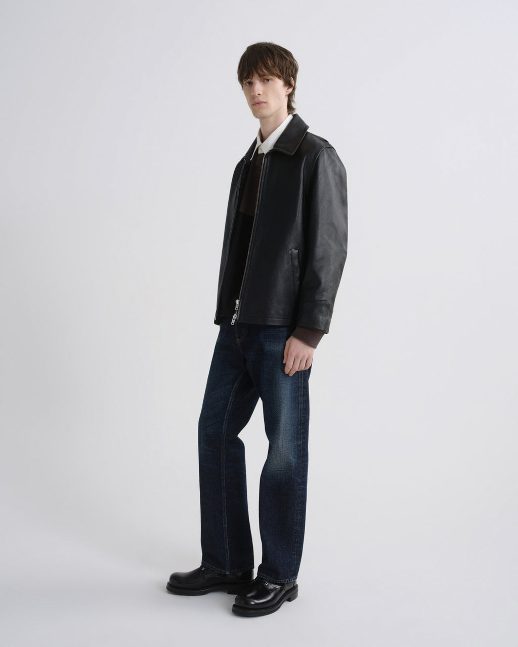 A model stands in a studio wearing a black leather jacket over a white collared shirt, a dark sweater, dark blue jeans, and black leather shoes, against a light gray background.