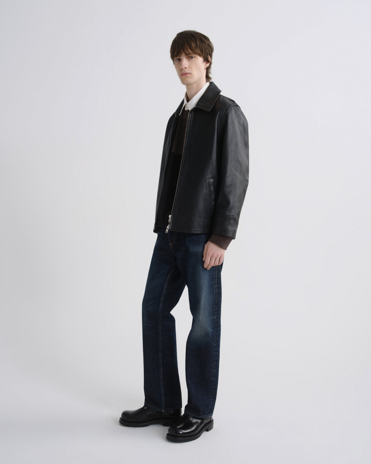 A model stands in a studio wearing a black leather jacket over a white collared shirt, a dark sweater, dark blue jeans, and black leather shoes, against a light gray background.