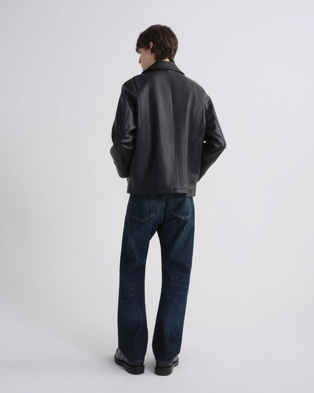 A man viewed from behind wearing a black leather jacket and dark blue jeans, standing against a light gray studio background.
