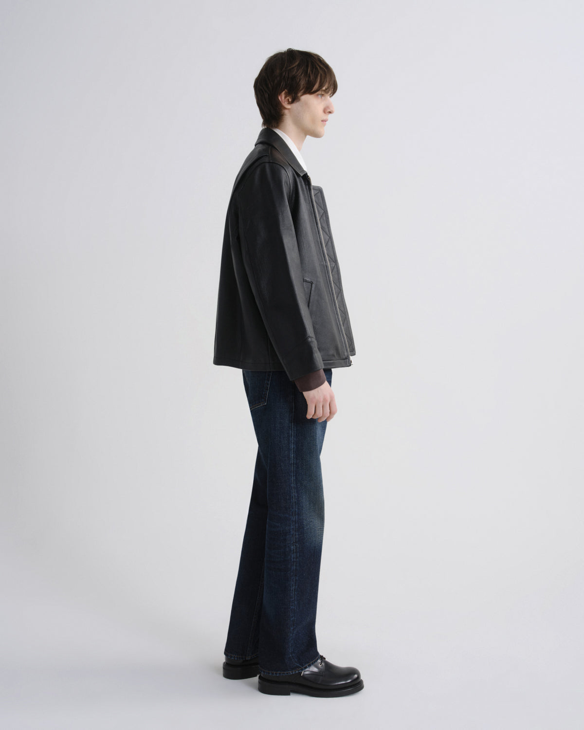 A man shown in profile wearing a black leather jacket, dark blue flared jeans, and black leather shoes against a light gray studio background.