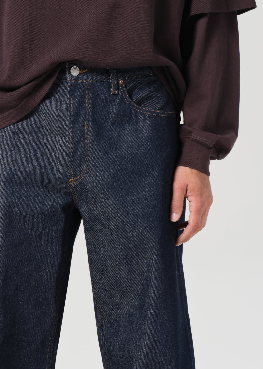 Person wearing dark blue jeans and a brown shirt on a light gray background