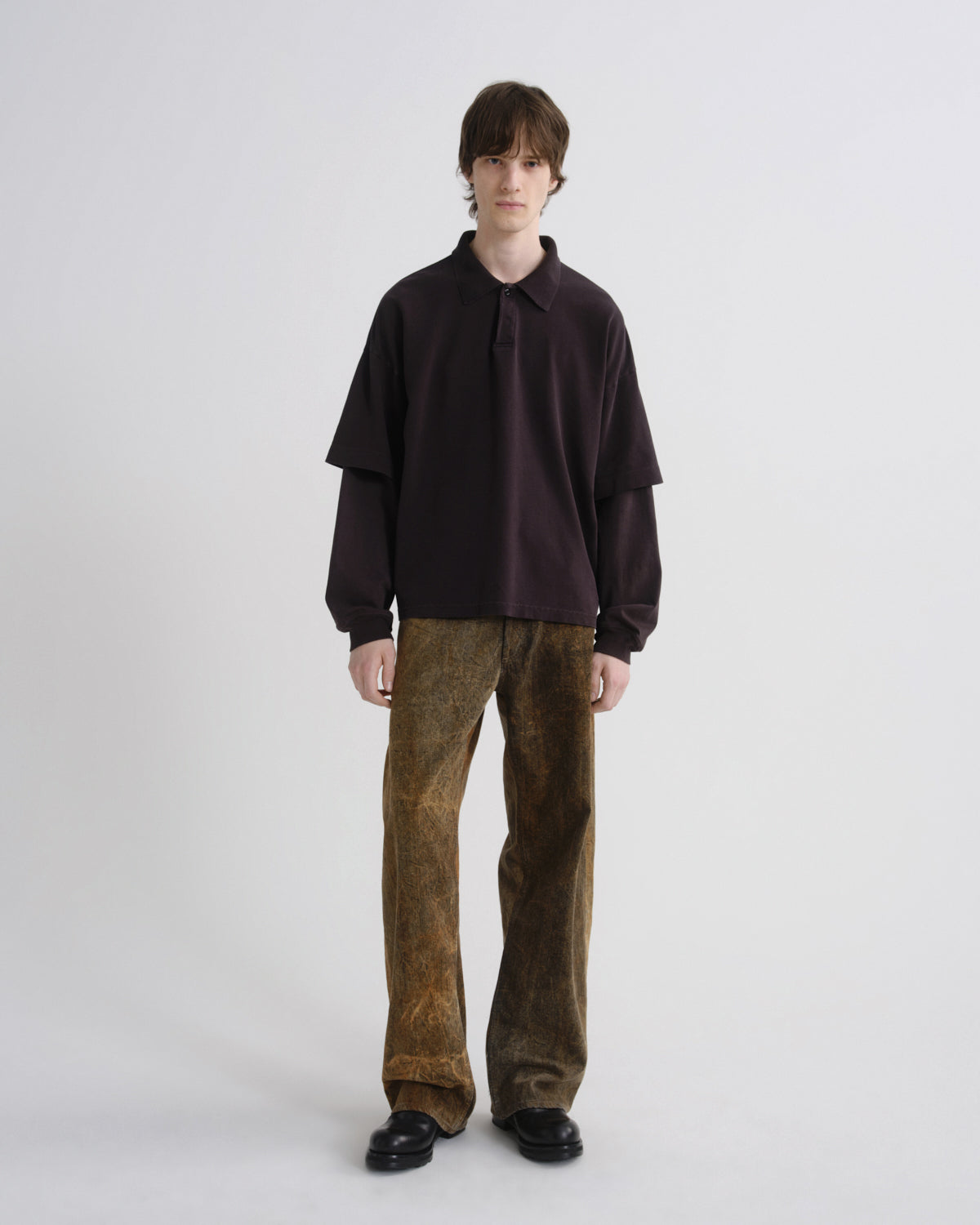 A model stands against a light gray studio background wearing a dark plum long-sleeve collared shirt with layered sleeves and brown textured wide-leg trousers.