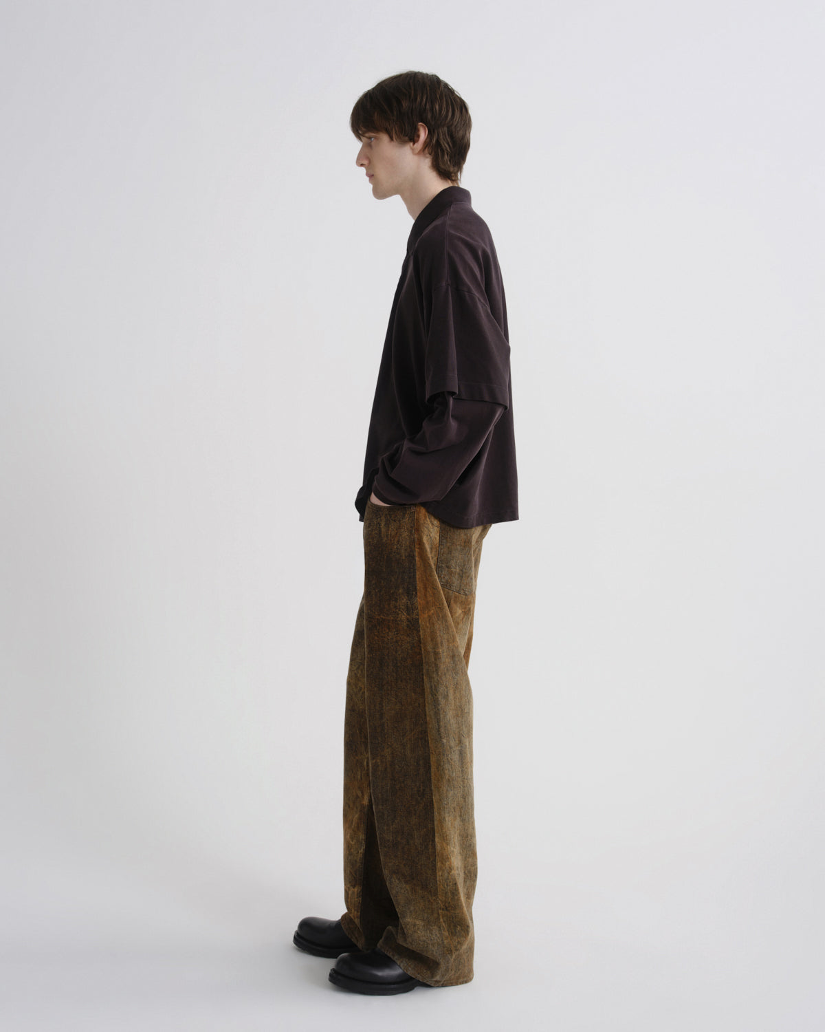 A model standing in profile wearing a dark brown long-sleeve shirt and wide-leg brown corduroy trousers against a plain light gray studio background.