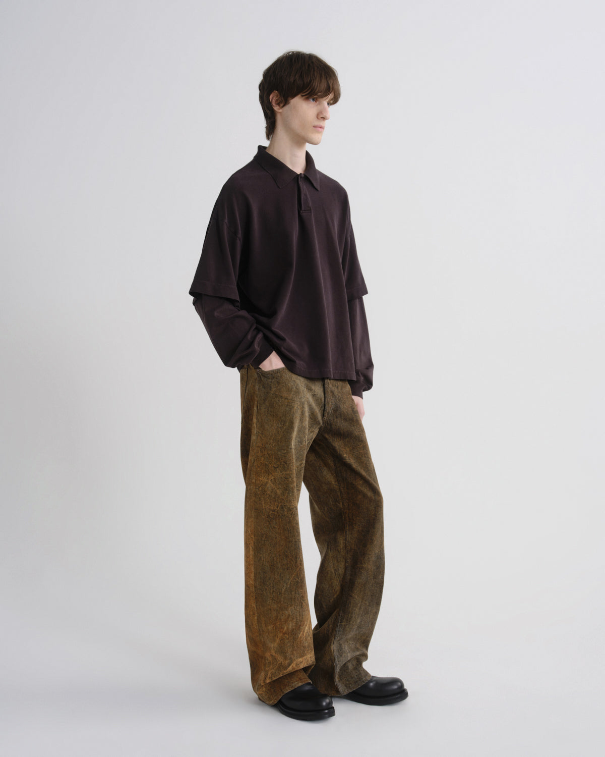 A model stands in profile wearing a dark plum long-sleeve button-up shirt and wide-leg brown corduroy trousers against a plain light gray studio background.