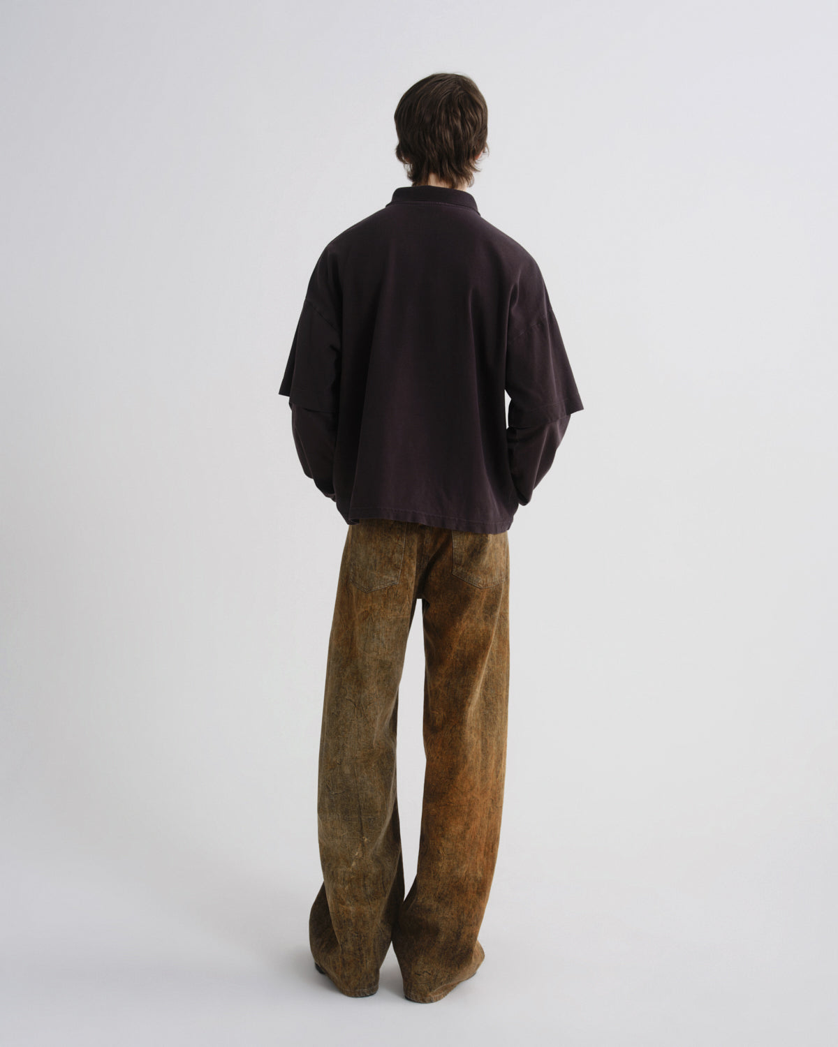 A model shown from the back wearing a dark plum-colored oversized shirt and brown wide-leg trousers, standing against a plain light gray background.