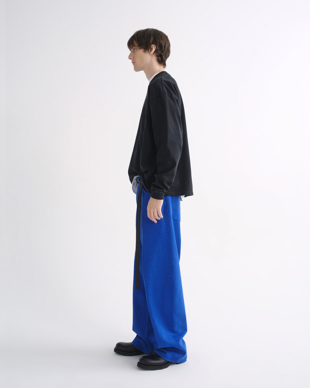Person wearing a black crewneck sweatshirt with bright blue pants and black shoes, side view, on a grey background