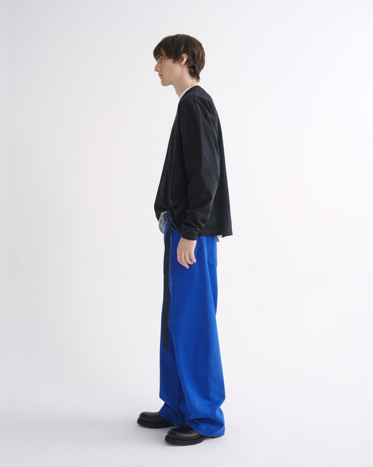 Person wearing a black crewneck sweatshirt with bright blue pants and black shoes, side view, on a grey background