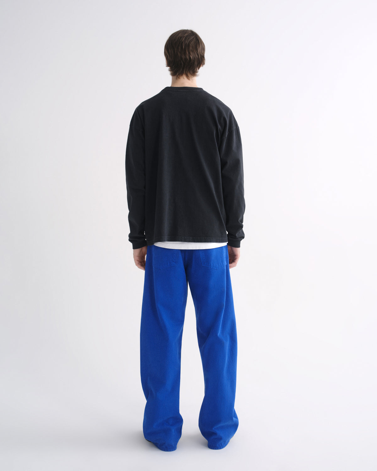 Person wearing a black crewneck sweatshirt with bright blue pants and black shoes, back view, on a grey background