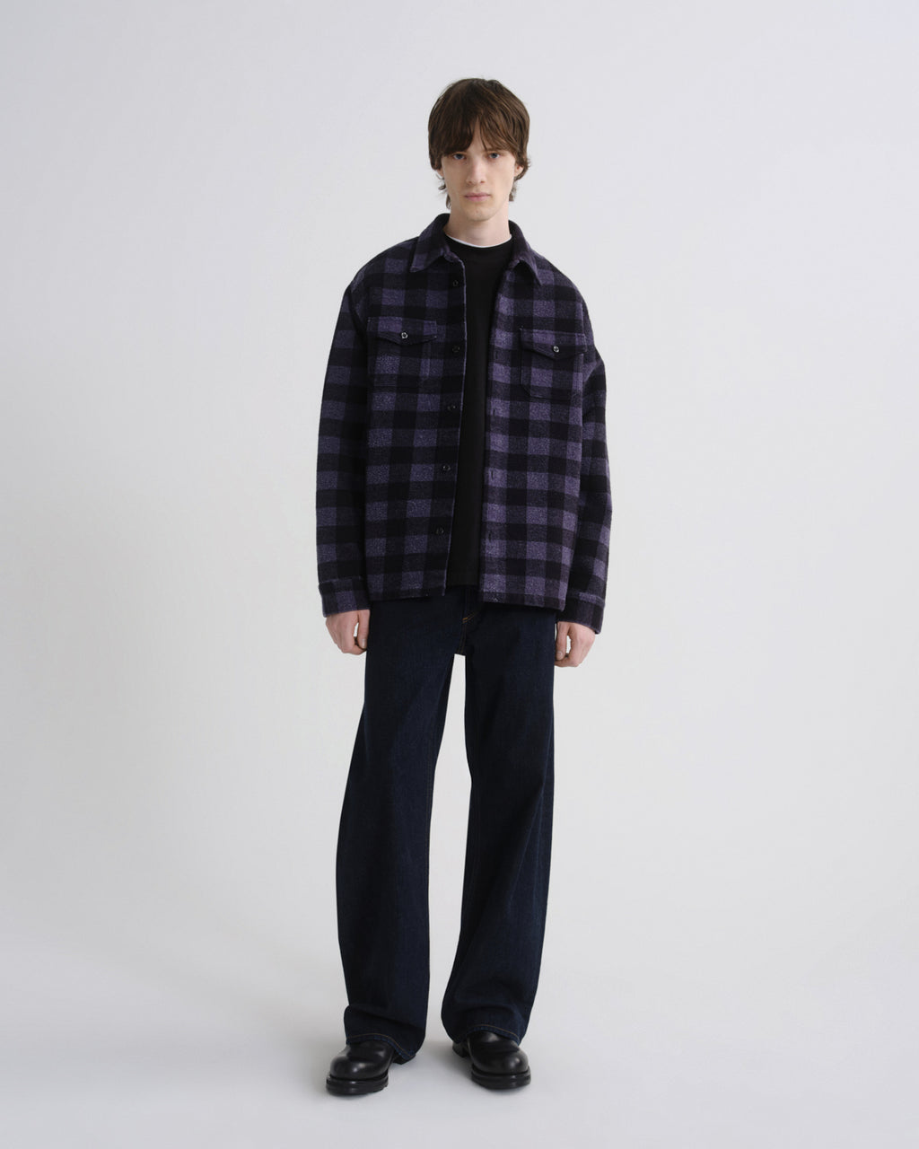 A model stands against a light gray studio background wearing a dark purple and black plaid overshirt over a black t-shirt, dark wide-leg jeans, and black leather shoes.