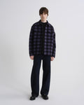 A model stands against a light gray studio background wearing a dark purple and black plaid overshirt over a black t-shirt, dark wide-leg jeans, and black leather shoes.