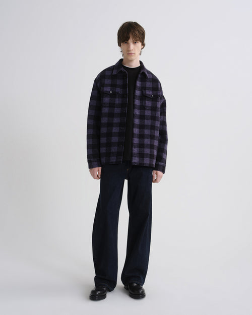 A model stands against a light gray studio background wearing a dark purple and black plaid overshirt over a black t-shirt, dark wide-leg jeans, and black leather shoes.