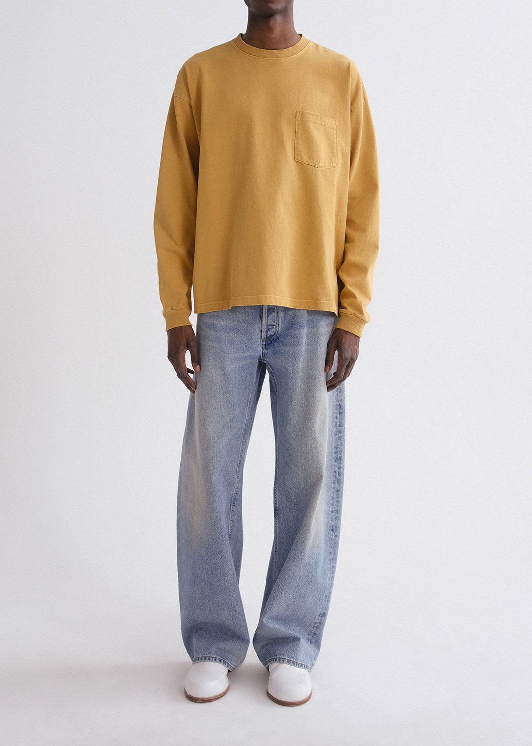 Person wearing a mustard yellow long-sleeve shirt and light blue jeans on a grey background