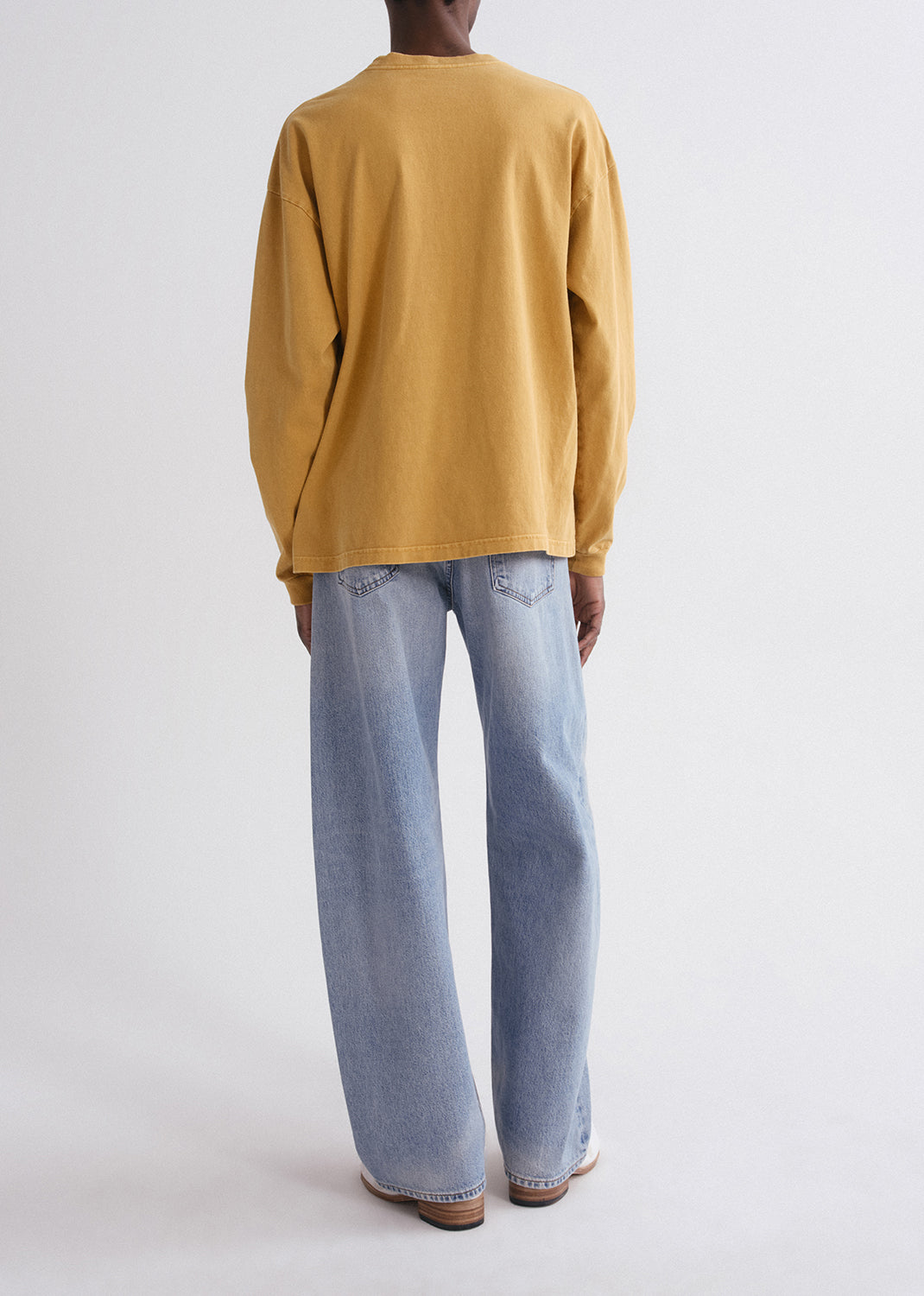 Person wearing a mustard yellow long-sleeve shirt and light blue jeans on a grey background