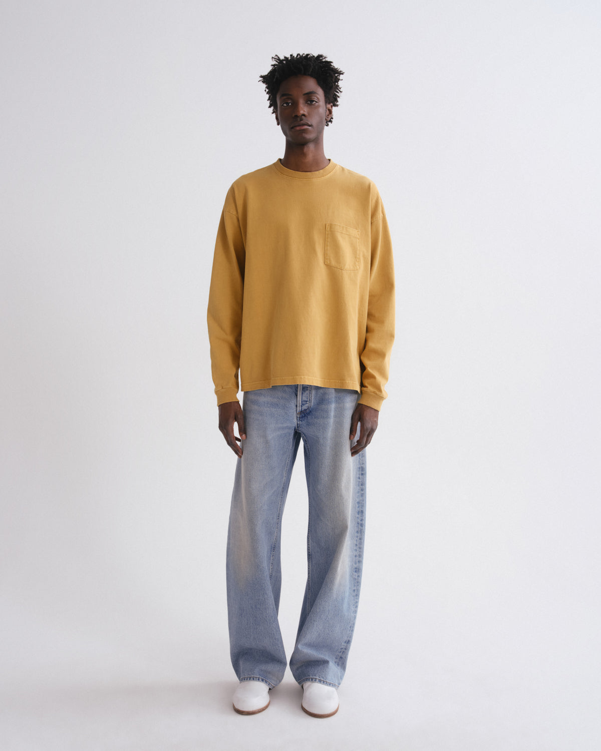 A model stands against a light gray studio background wearing a mustard yellow long-sleeve crewneck sweatshirt with a chest pocket, light-wash wide-leg jeans, and white sneakers.