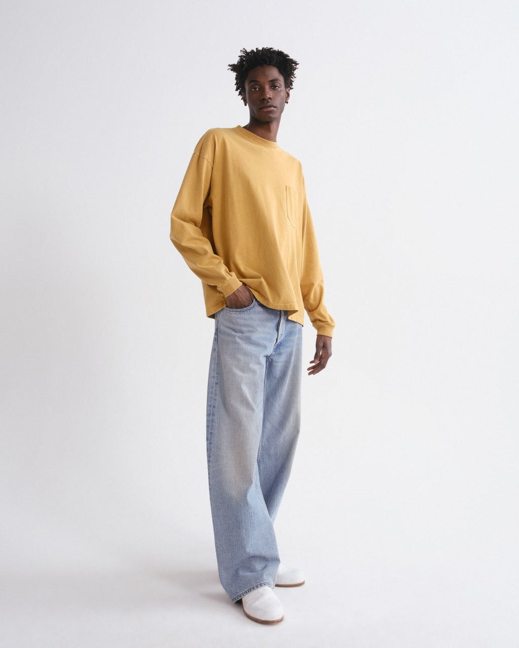 A model stands against a plain white studio background wearing a mustard yellow long-sleeve crewneck sweatshirt, light-wash wide-leg jeans, and white sneakers.