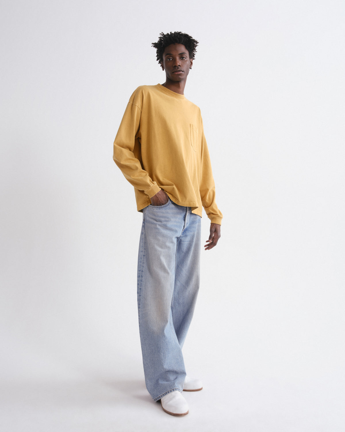 A model stands against a plain white studio background wearing a mustard yellow long-sleeve crewneck sweatshirt, light-wash wide-leg jeans, and white sneakers.