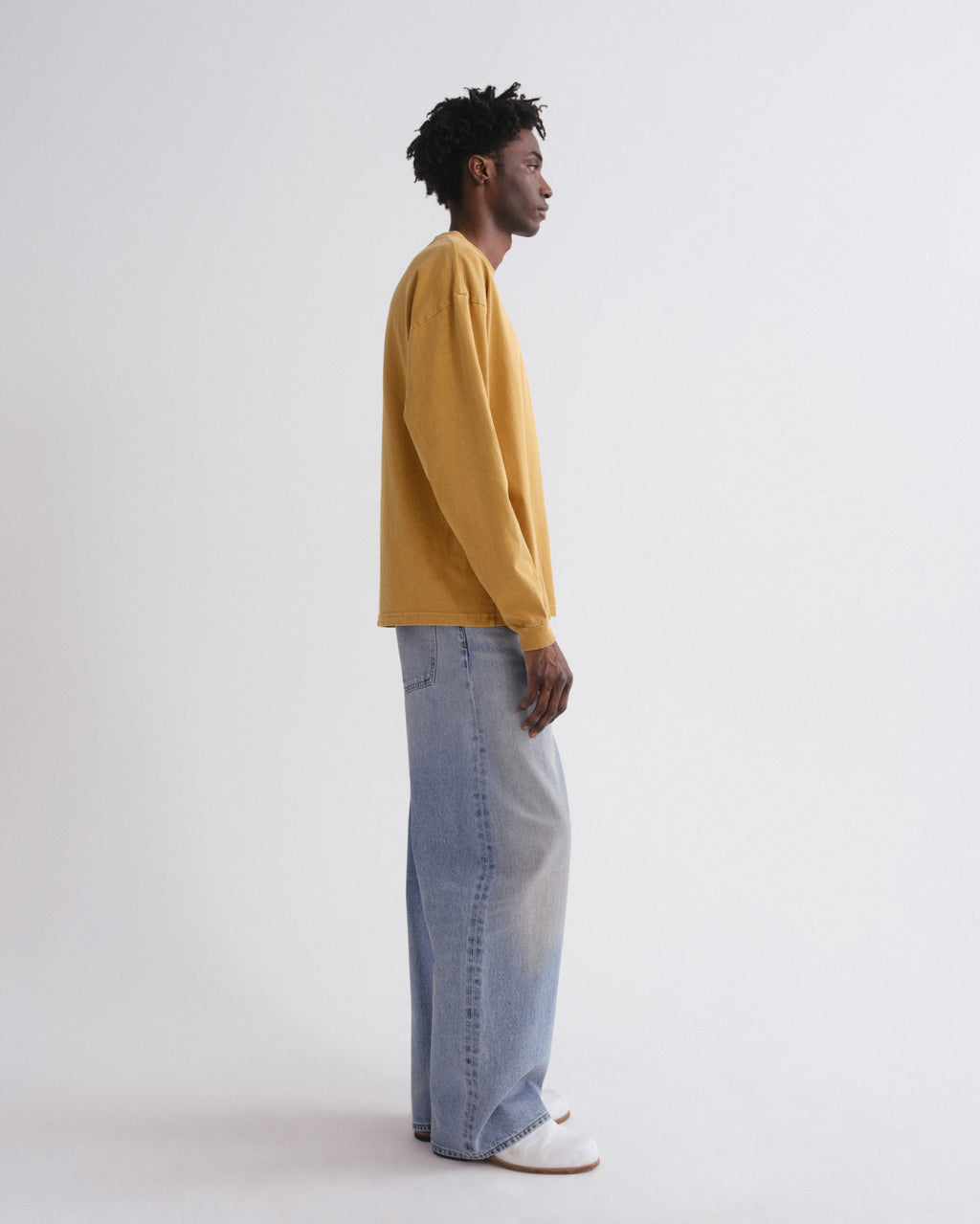 A man in profile wearing a mustard yellow long-sleeve sweatshirt, light-wash wide-leg jeans, and white slip-on shoes, standing against a plain light gray studio background.