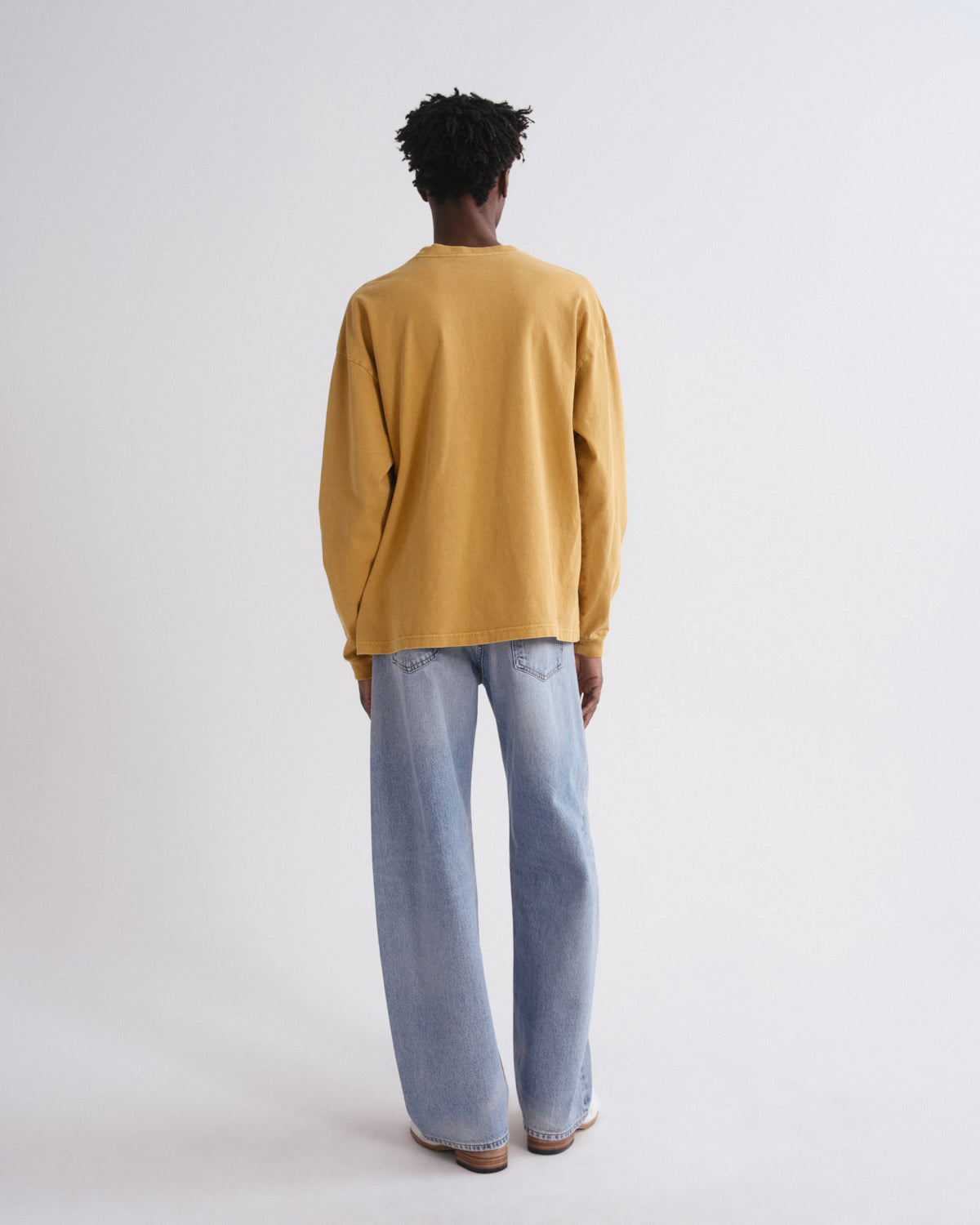 A person shown from the back wearing a mustard yellow long-sleeve top and light blue wide-leg jeans against a plain light gray studio background.