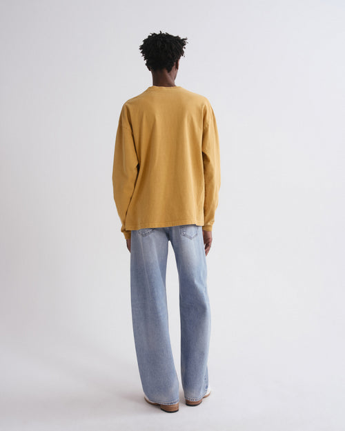 A person shown from the back wearing a mustard yellow long-sleeve top and light blue wide-leg jeans against a plain light gray studio background.