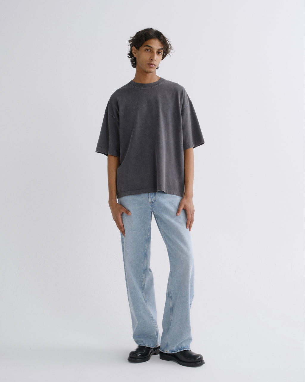 A model stands against a light gray studio background wearing an oversized dark gray t-shirt, light blue wide-leg jeans, and black leather shoes.