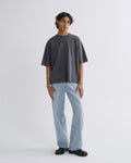 A model stands against a light gray studio background wearing an oversized dark gray t-shirt, light blue wide-leg jeans, and black leather shoes.