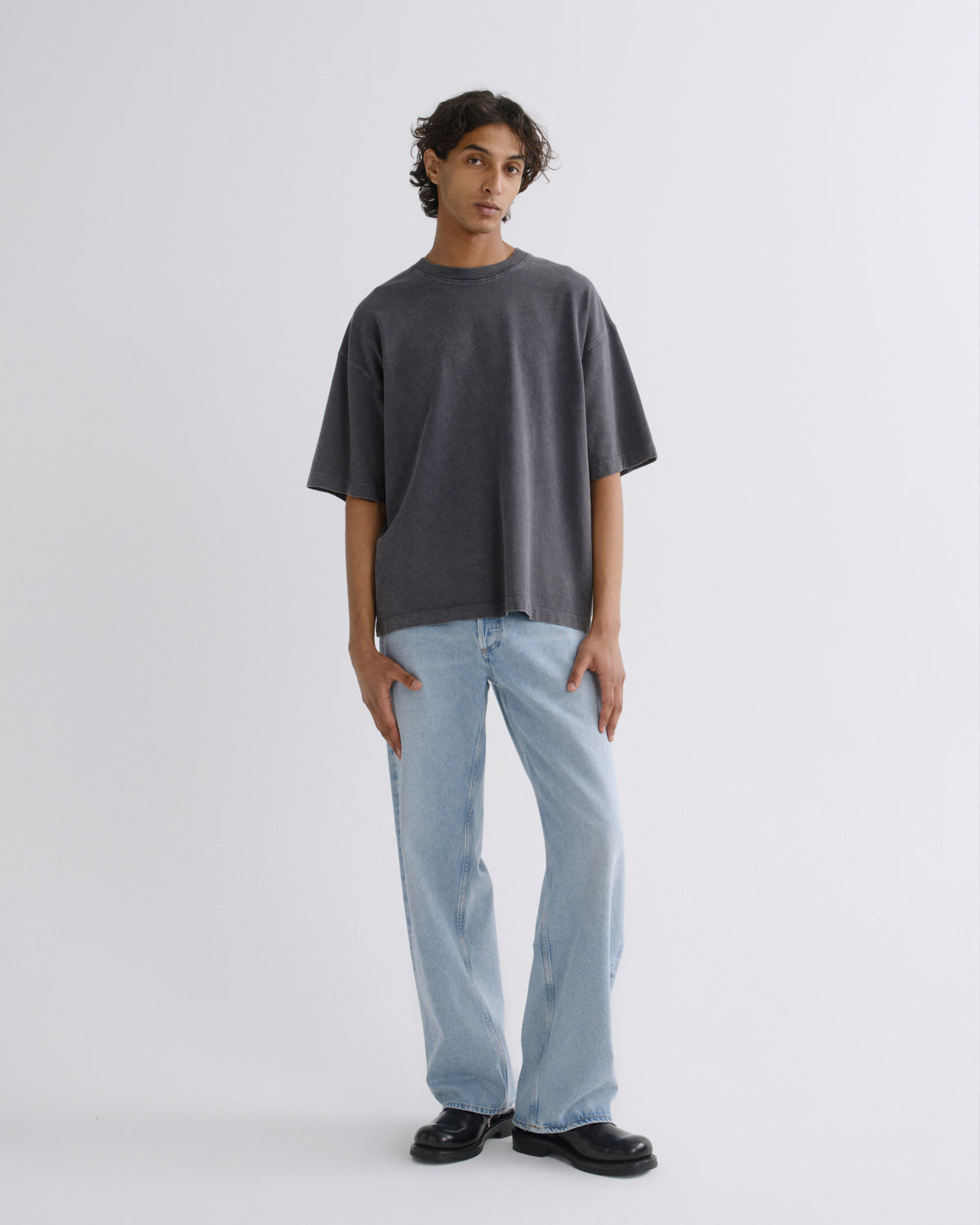 A model stands against a light gray studio background wearing an oversized dark gray t-shirt, light blue wide-leg jeans, and black leather shoes.