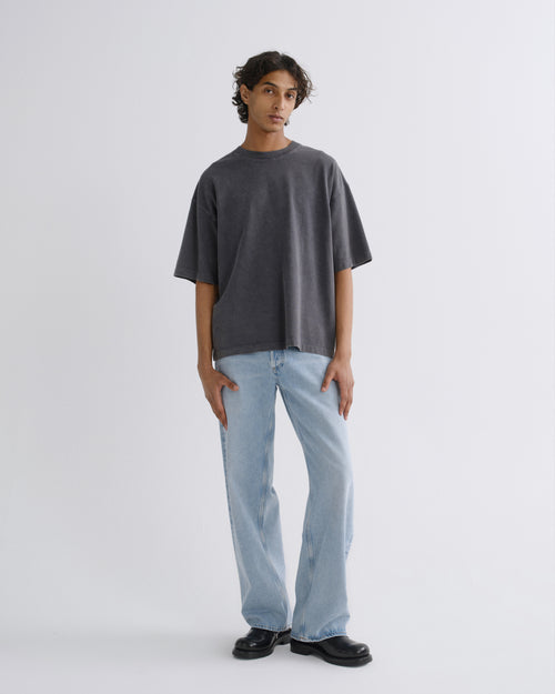 A model stands against a light gray studio background wearing an oversized dark gray t-shirt, light blue wide-leg jeans, and black leather shoes.