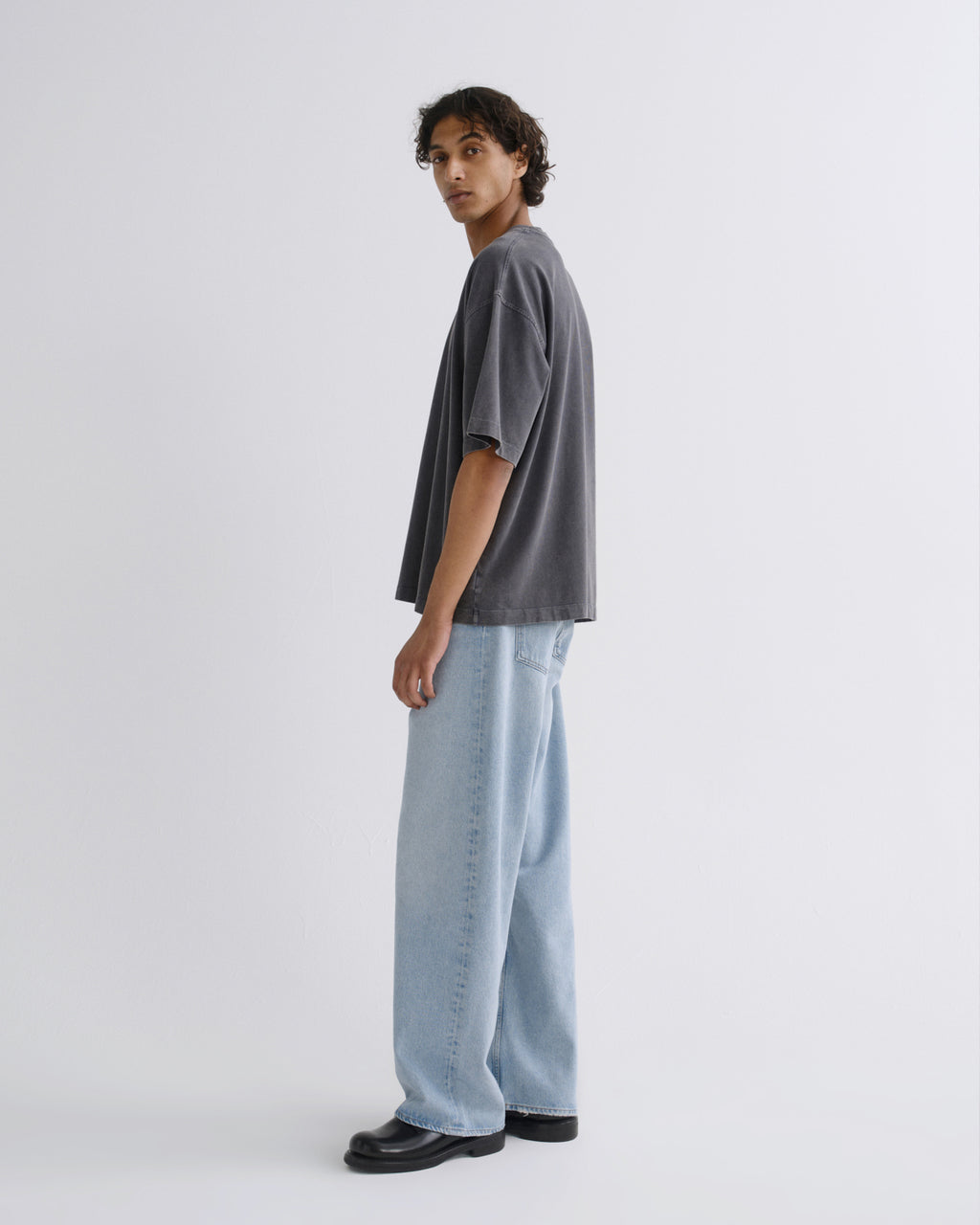 A model wearing a dark gray oversized t-shirt, light blue wide-leg jeans, and black leather shoes, standing in profile against a light gray studio background.