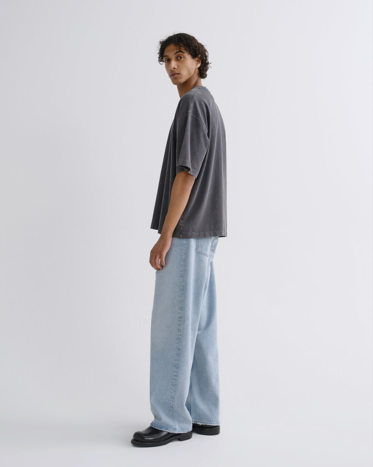 A model wearing a dark gray oversized t-shirt, light blue wide-leg jeans, and black leather shoes, standing in profile against a light gray studio background.