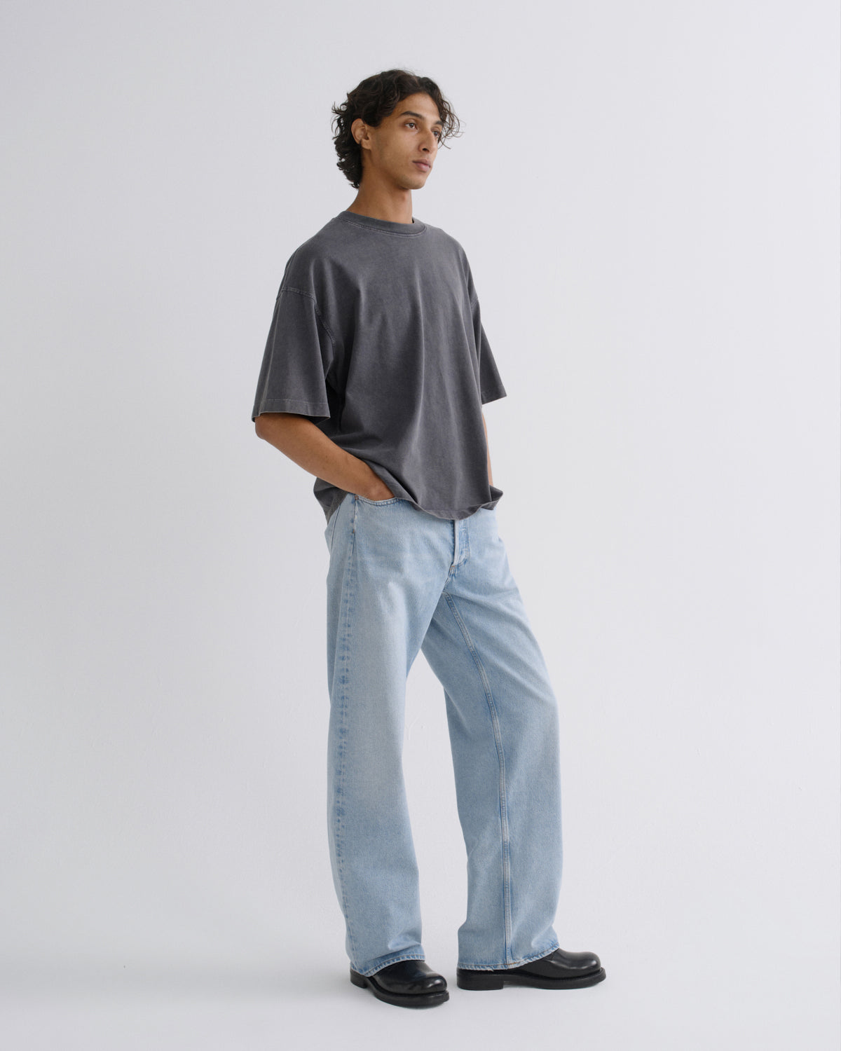 A man modeling an oversized dark gray t-shirt and light-wash wide-leg jeans against a plain light gray studio background.