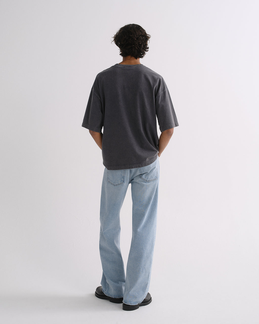 A man viewed from behind wearing an oversized dark charcoal gray t-shirt and light blue straight-leg jeans, standing against a plain light gray studio background.