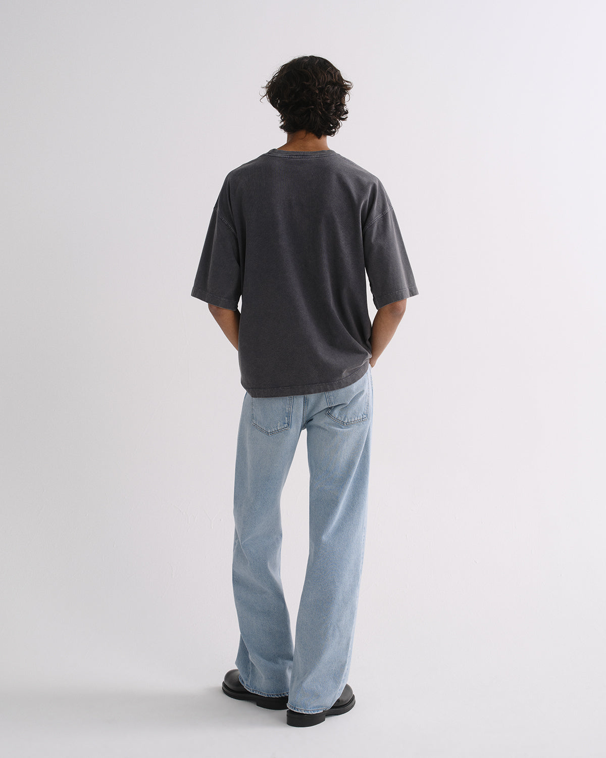 A man viewed from behind wearing an oversized dark charcoal gray t-shirt and light blue straight-leg jeans, standing against a plain light gray studio background.