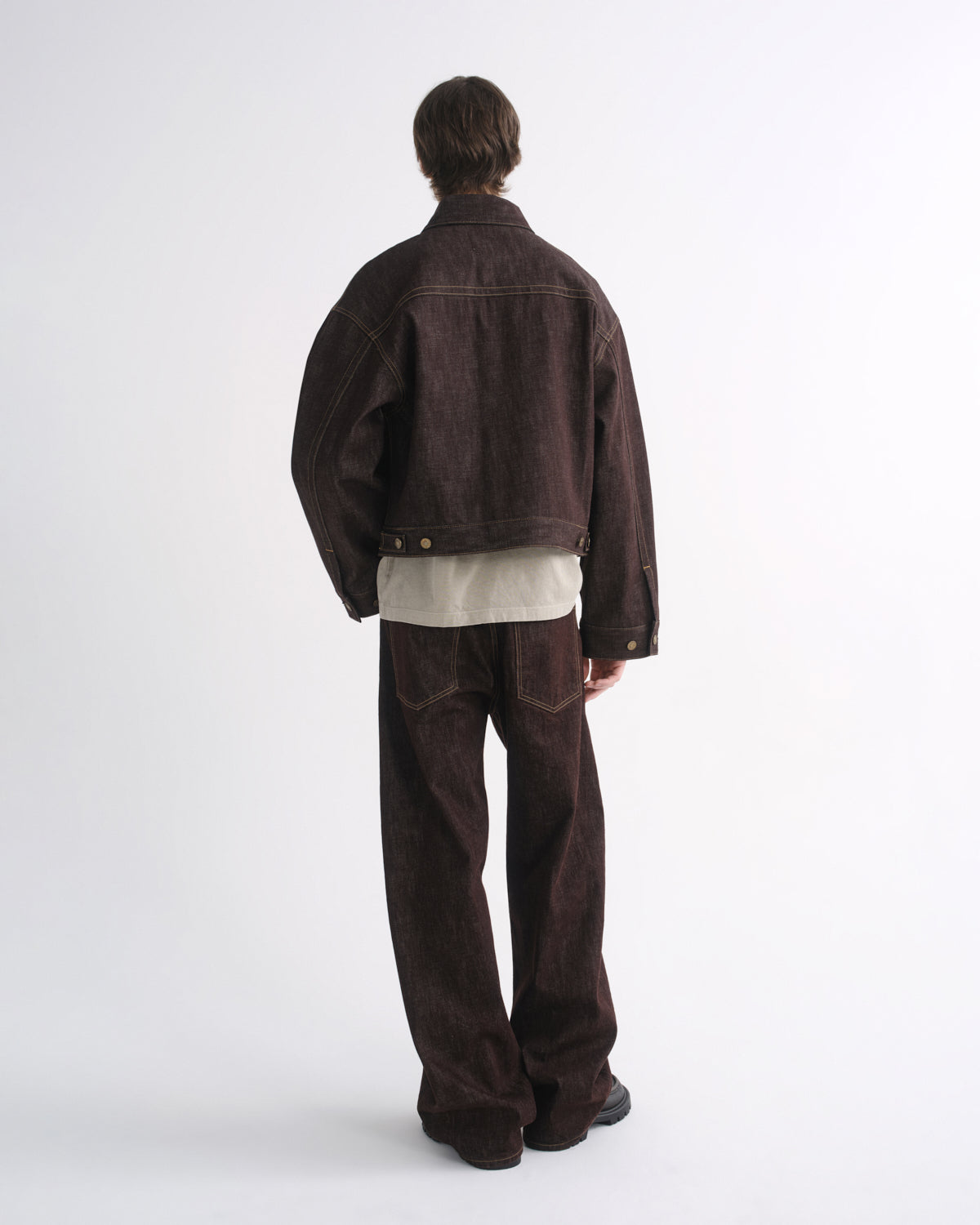 Person wearing a dark brown denim jacket with matching dark brown jeans and black shoes, back view, on a grey background