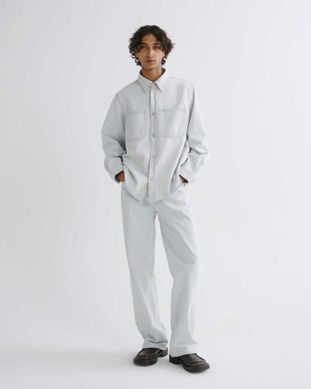 A model stands against a light gray studio background wearing a light gray long-sleeve button-up shirt and matching light gray trousers, paired with dark brown loafers.