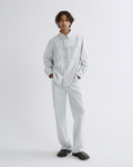 A model stands against a light gray studio background wearing a light gray long-sleeve button-up shirt and matching light gray trousers, paired with dark brown loafers.