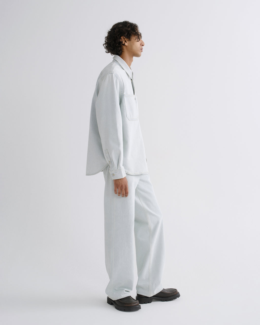 A model shown in profile wearing a light blue oversized button-up shirt and matching wide-leg trousers, paired with dark brown shoes, against a plain light gray studio background.