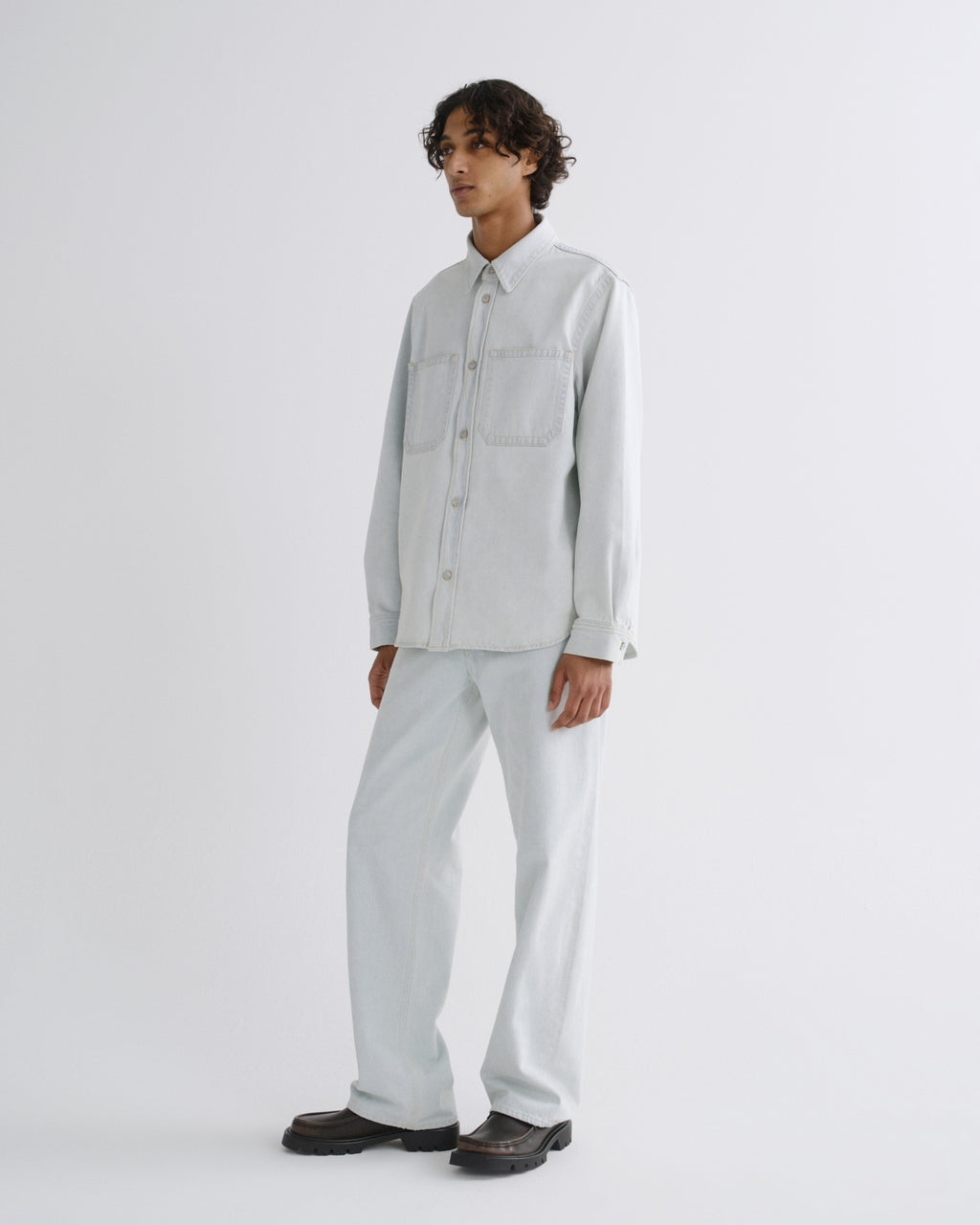 A model stands in a studio wearing a light gray long-sleeve button-up shirt and matching light gray trousers, paired with dark brown shoes, against a plain light gray background.
