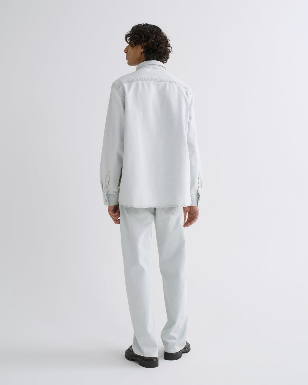 A model shown from the back wearing a light gray long-sleeve button-up shirt and matching light gray trousers, paired with black shoes, against a plain light gray studio background.